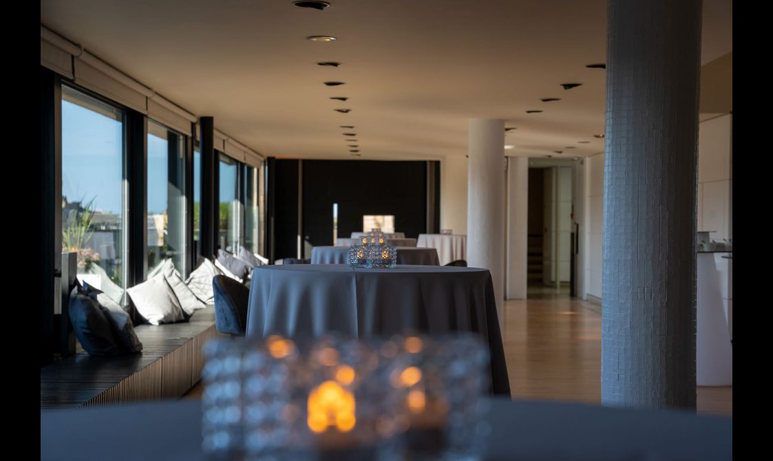 A long room with tables set out. The left hand wall is lined with windows look out onto Edinburgh. There are cushions by the windows for sitting.