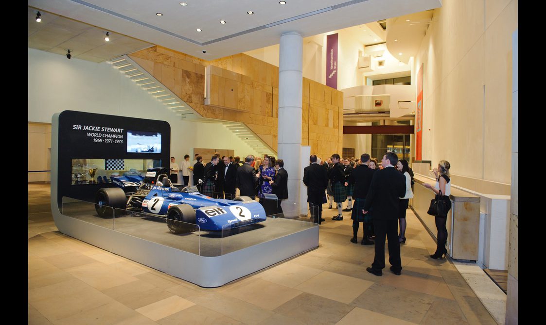 A group of people at a drinks reception in a large museum space with stone floors, high ceilings, and white walls. There is a formula one racing car on a platform.