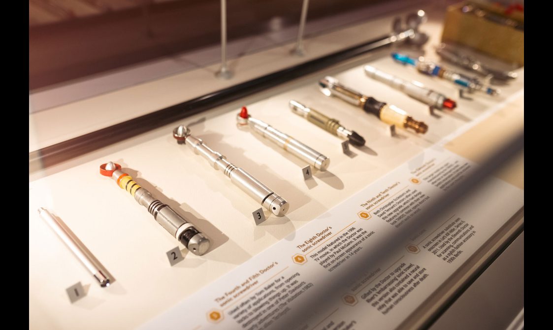 A series of Sonic Screwdrivers from Doctor Who are laid out in bright white glass display case.