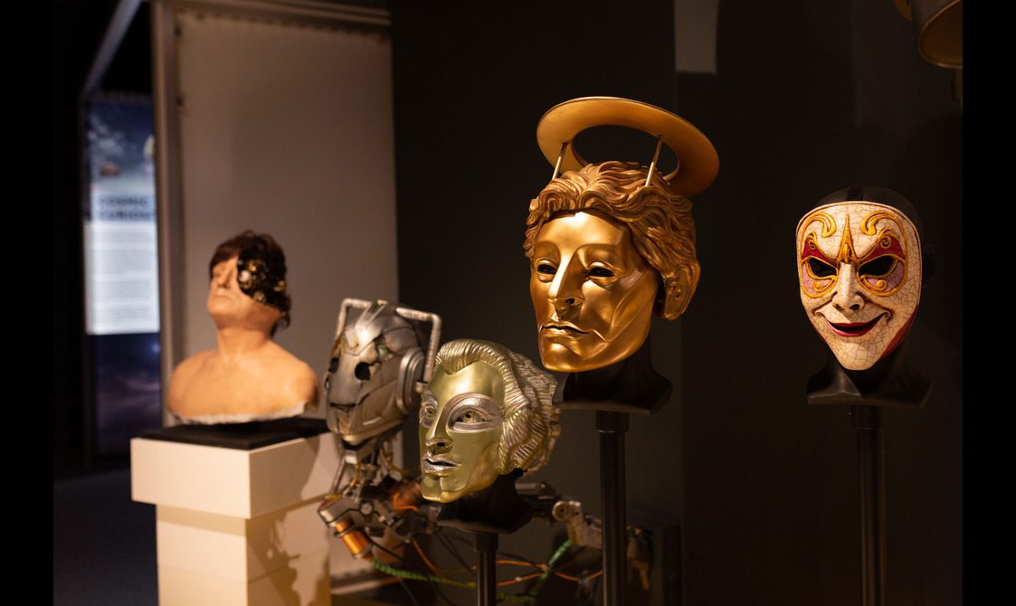 A lineup of five disembodied robot or android heads from Doctor Who in a futuristic museum gallery.