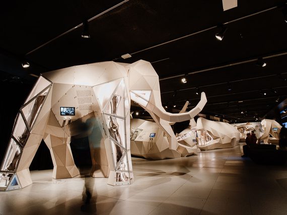 Large 3D origami like sculptures of giant animals, in the giants exhibition space. In the foreground is a woolly mammoth, with glass panels on the legs that show real fossilised bones inside.
