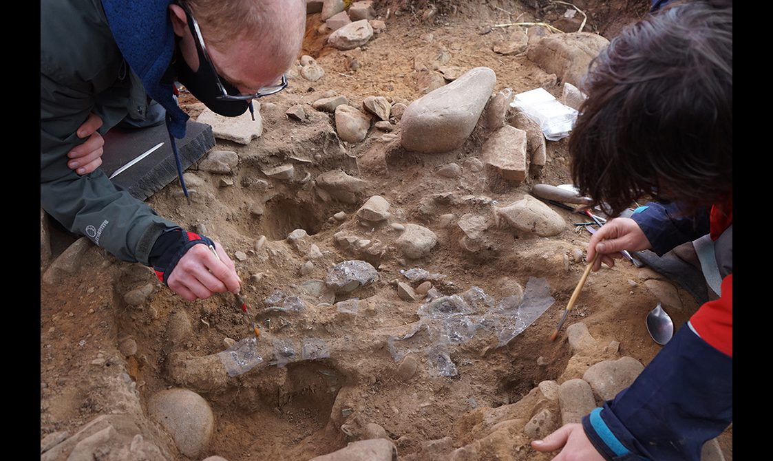 Two curators carefully brushing away surface dirt from objects in the ground