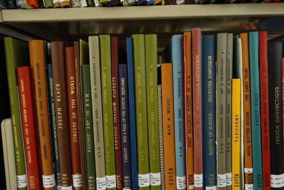 A shelf of thin, hardcover books with different coloured spines. They have library labels at the base.