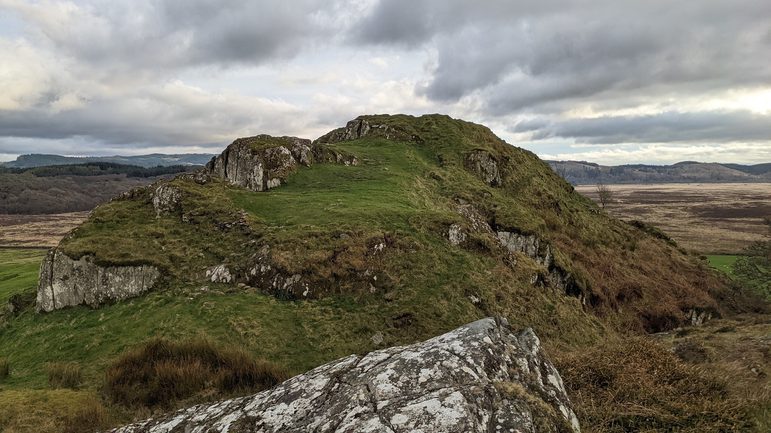 A grassy hill with rocks and heather. More hills and forests are visible behind the hill.