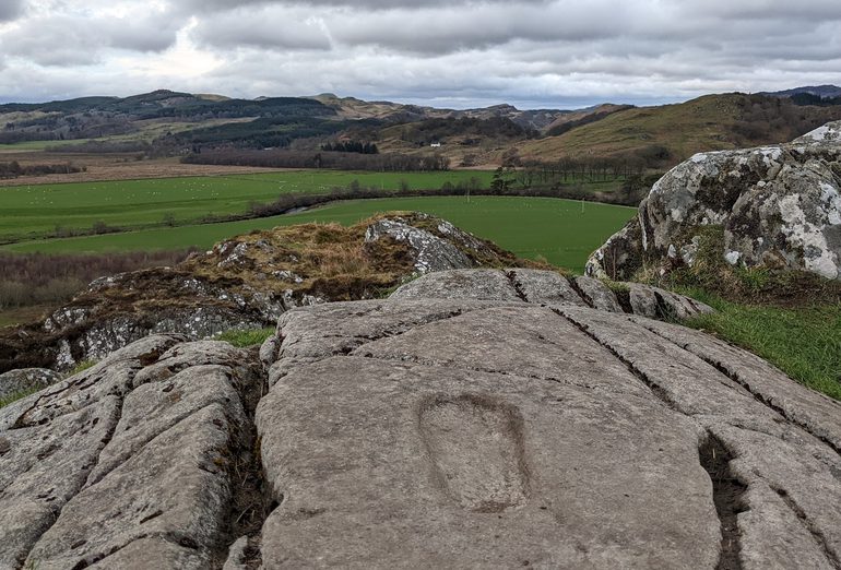 A rock with a footprint imprinted on its surface overlooking green fields and green and brown hills
