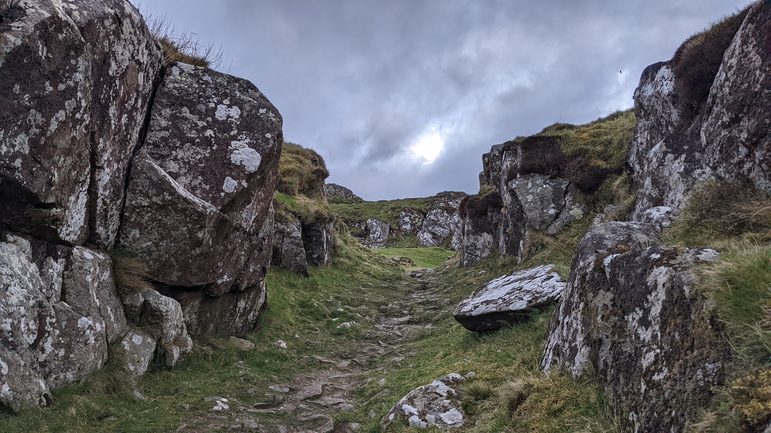 A narrow stone lined path with large rocks on either side with patches of grass