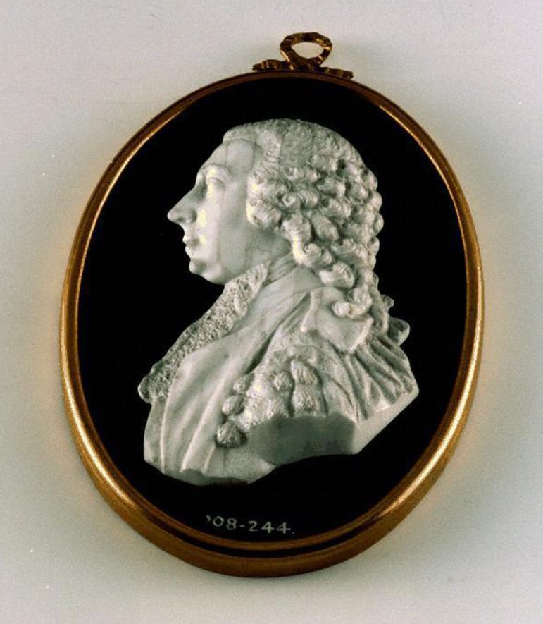 An oval medallion with a gold frame and black background with a white raised portrait of a man with long curly hair wearing a ruffled high neck shirt.