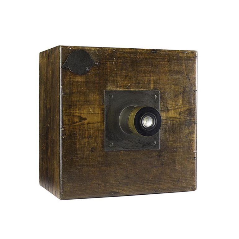 An early camera made of a square wooden box with a brass plate in the centre with a round lens protruding.