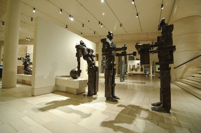 A bright white gallery space with stone floors and track lighting on the ceiling. There are three large, blocky sculptures of people.