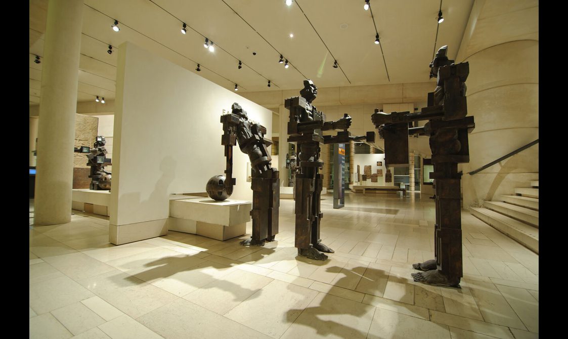 A bright white gallery space with stone floors and track lighting on the ceiling. There are three large, blocky sculptures of people.