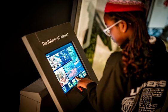 A child with braids and glasses looks at a touchscreen with the title 'The Habitats of Scotland'.