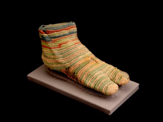 A faded striped knitted sock with a split toe design and various holes on display on a mount.