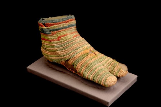 A faded striped knitted sock with a split toe design and various holes on display on a mount.