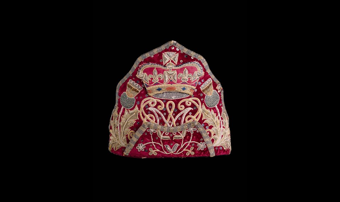 Red triangle shaped cap decorated with yellow and green embroidery of two thistles in opposite corners, 'A' and 'M' initials in between the thistles, and a crown placed atop the initials.