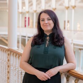 Headshot of Emily Brown from the waist up, wearing business casual clothing and smiling. She is leaning on a railing overlooking National Museums Scotland’s Grand Gallery.