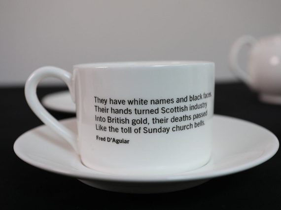 A white porcelain teacup with text written on it placed on a saucer set against blurry background.