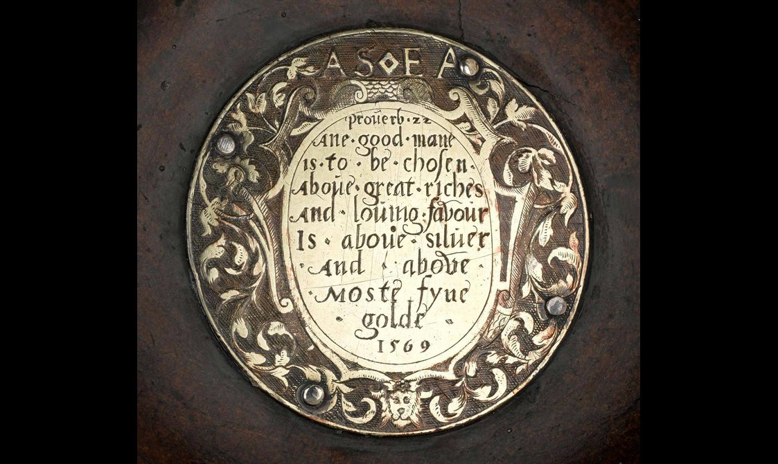 A round plaque with floral detail around the border with text engraved in the middle and the year 1569