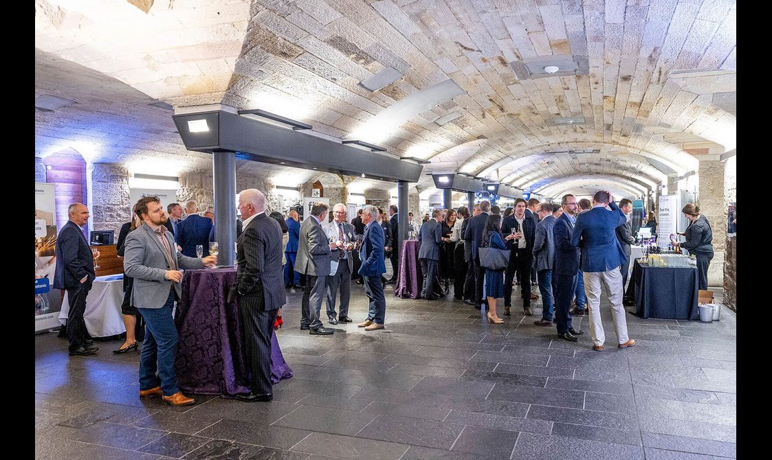 People at a drinks reception in a long hall with stone floors and a low arched ceiling.