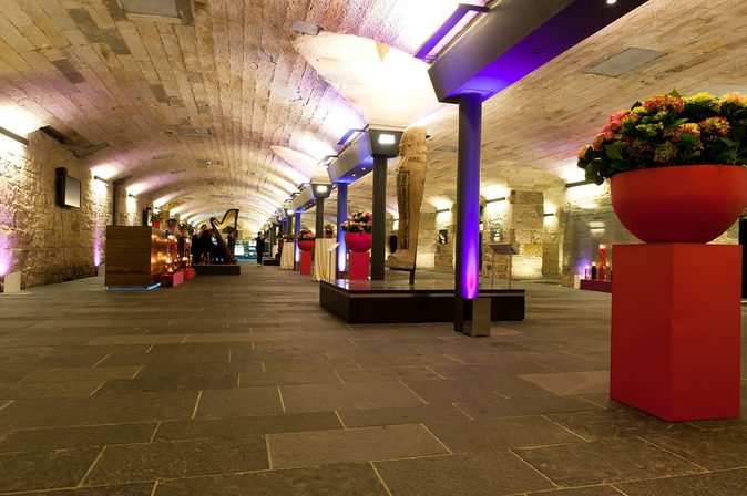 A long hall with stone floors and a low arched ceiling. There are supporting pillars lining the length of the hall.