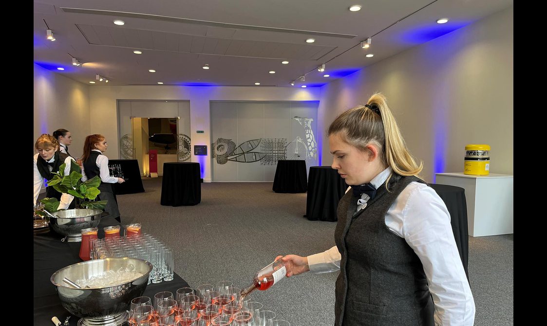 Catering staff in tweed waistcoats setting up an event space for a drinks reception.