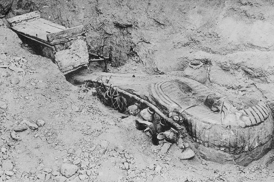 A black and white image of an ancient Egyptian coffin half buried in the soil