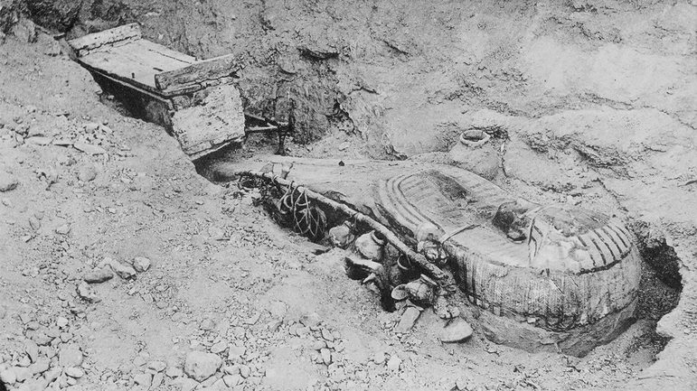 A black and white image of an ancient Egyptian coffin half buried in the soil