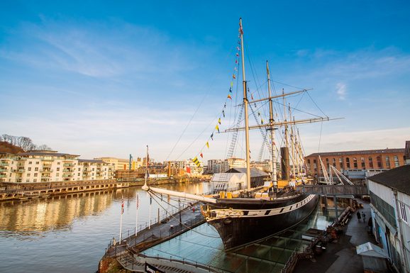 Old fashioned ship with masts on a platform beside a river with residential buildings on either side.
