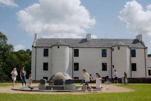 Low and long white building with two small conical turrets and a fountain sculpture of the earth. Sunny day with people playing happily on the grass.