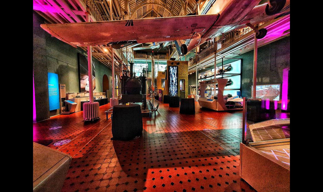 A multi-level museum gallery colourfully lit up for an evening event. There are different aircraft suspended from the ceiling, and different vehicles around the gallery.