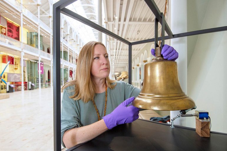 A curator hanging a brass bell on a frame in a large museum gallery.