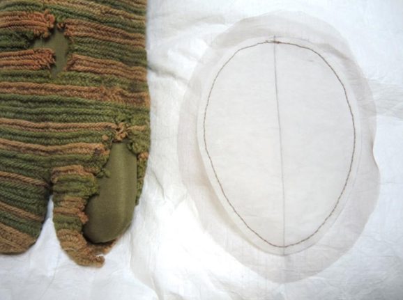 A faded striped knitted sock with a split toe design and various holes sits beside a thin piece of transparent fabric cut in an oval shape.