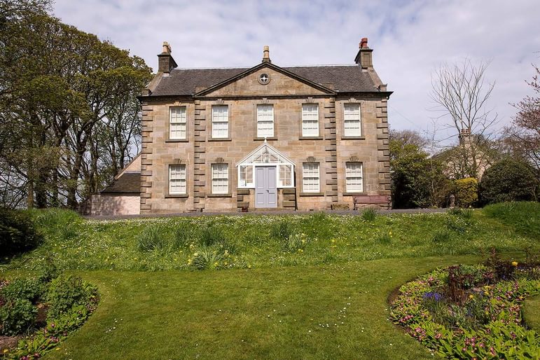 A large two story stone house with a green lawn with flower beds in front.