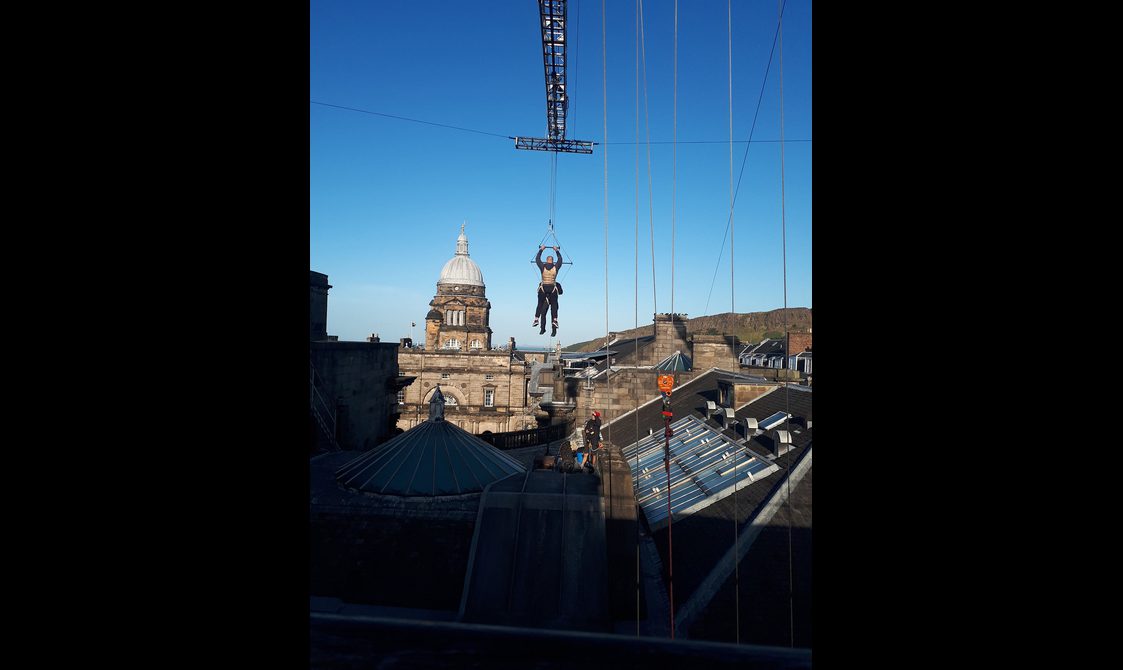 Two stunt people hang from a crane above the rooftop of the National Museum of Scotland against a clear blue sky.