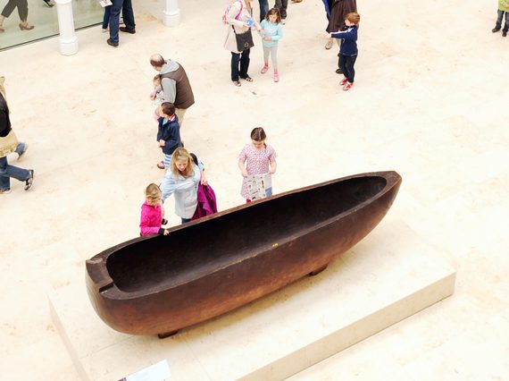 A large oval shaped wooden object with a hollowed out middle on display in a museum with people looking at it