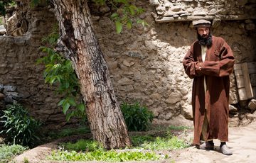 A man in a brown full-length coat stands next to a tree trunk in front of a stone wall.