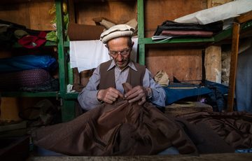 A man in a white flat hat stands in front of shelves holding different fabrics. He is hand-stitching a brown garment.