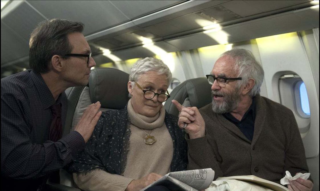 An older woman and man sit in airplane seats. The man points at another man crouching down in the aisle.