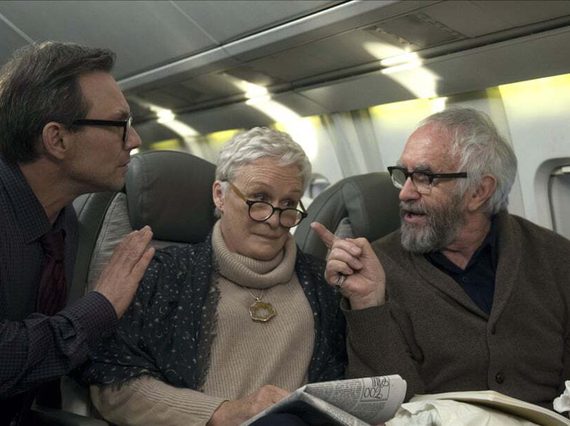 An older woman and man sit in airplane seats. The man points at another man crouching down in the aisle.