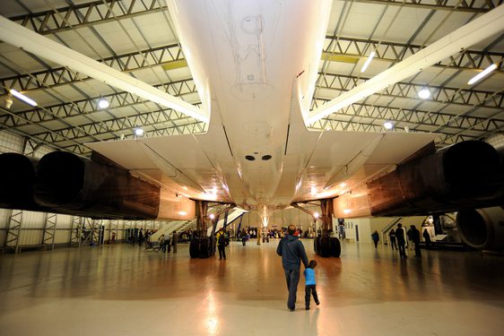An adult and child stand beneath the tail end wings of a Concorde aircraft stationed inside a large hangar.