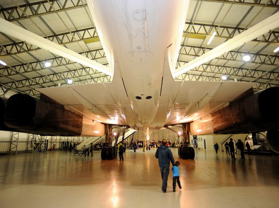 An adult and child stand beneath the tail end wings of a Concorde aircraft stationed inside a large hangar.