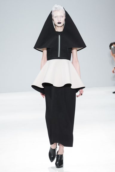 A model with short white hair and black lips wearing a black and white dress, walking down the runway. The dress features a black hood and she is wearing black heeled shoes.