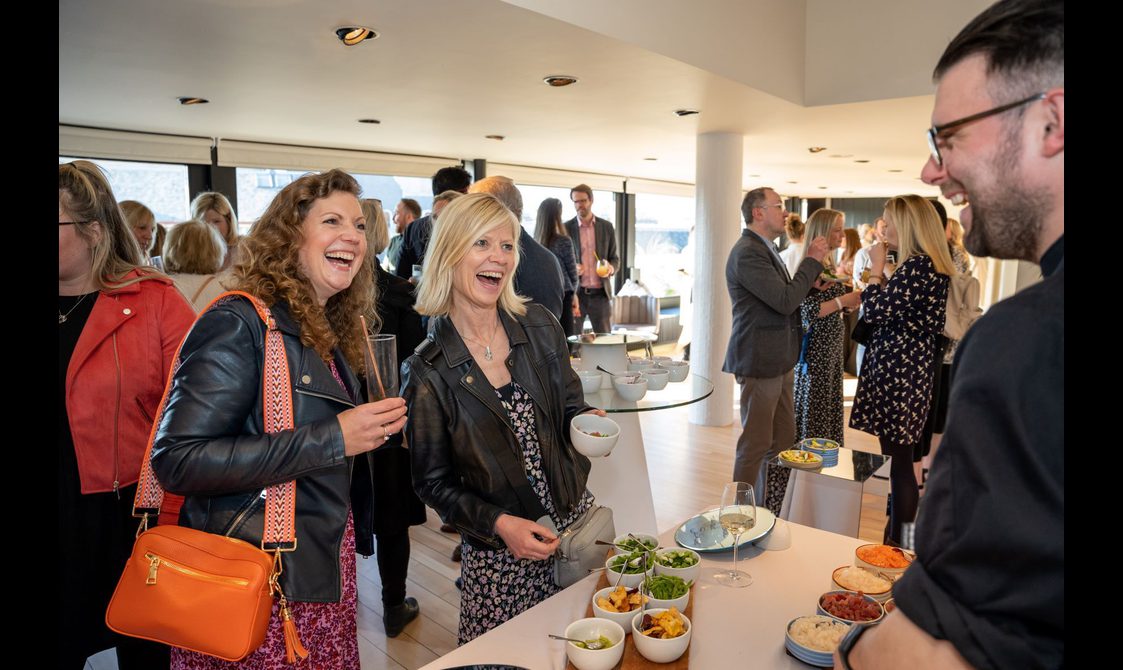 Two women standing holding drinks and snacks smile at a man behind a table full of food. They are in a crowded room with windows at the back.