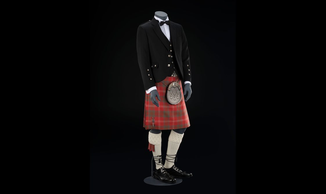 A mannequin wearing a formal outfit of a red kilt and white shirt with a black waistcoat, jacket, and bow tie.