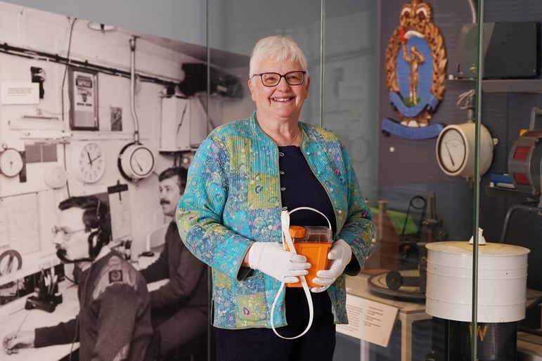 A woman wearing a colourful jacket is standing, smiling at the camera, holding an orange box and wearing white gloves, whilst standing in the cold war scotland exhibition space.