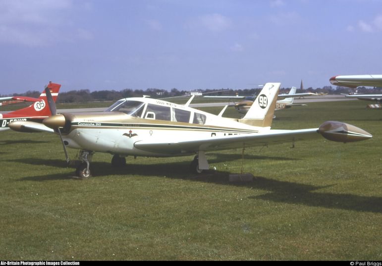 A Piper PA-24 'Comanche' 260B aircraft parked on a grassy airfield. The aircraft is white with gold and black stripes along the body.