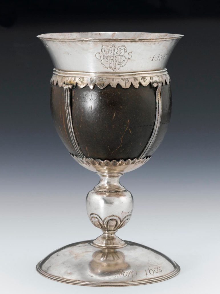 A colour photograph of a cup made from coconut against a grey gradient background. The cup has a splayed silver rim at the top and a tall silver stem. The base is engraved with the date 1608 and the rim has a coat of arms with the letters G and S and the date 1588.