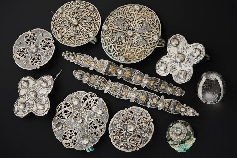 Cluster of silver objects on a black surface viewed from above. Two decorated metal strips in the centre with 9 brooches, mostly round, surrounding them.