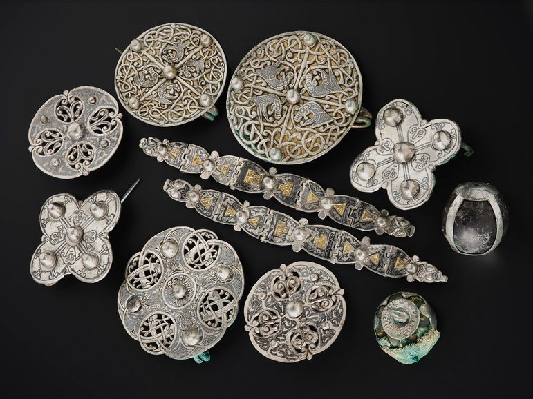 Cluster of silver objects on a black surface viewed from above. Two decorated metal strips in the centre with 9 brooches, mostly round, surrounding them.