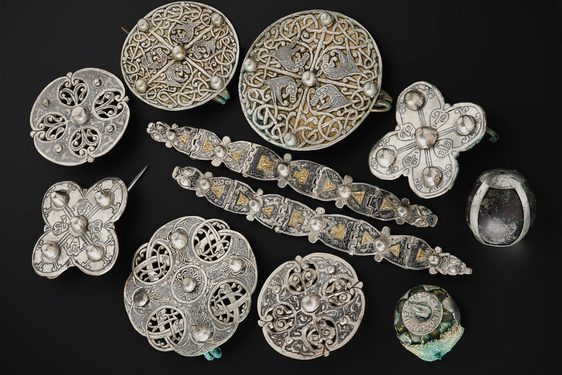 Cluster of silver objects on a black surface viewed from above. Two decorated metal strips in the centre with 9 brooches, mostly round, surrounding them.