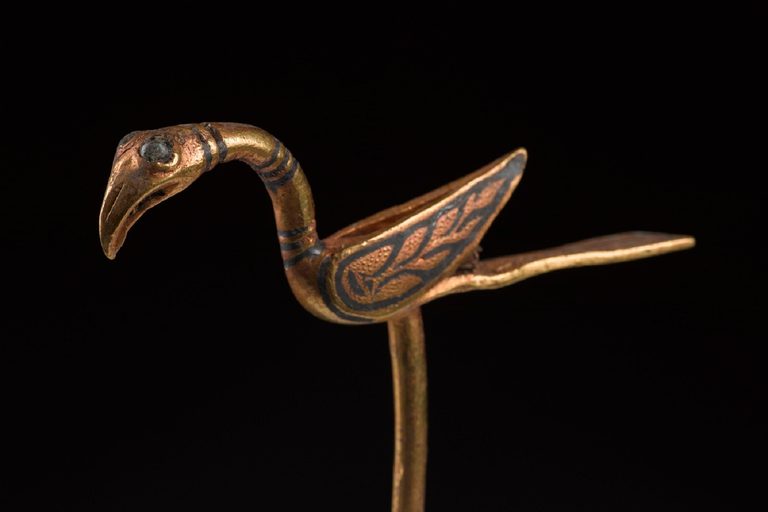 Golden pin shaped like a bird, with long, flat tail, upturned wings and flamingo-like beak.